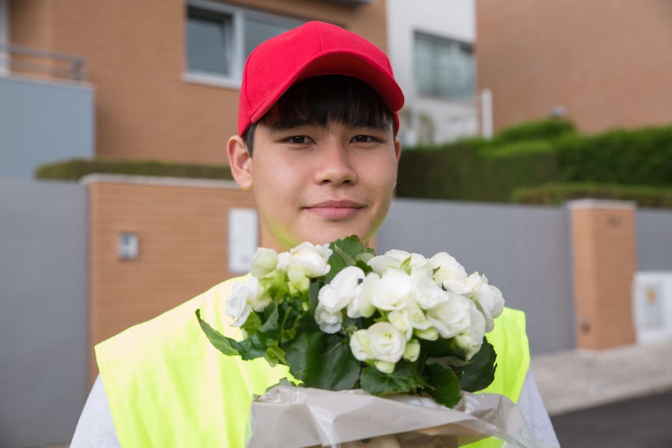 A young male delivery person with short dark hair, wearing a red cap and a high-visibility yellow vest, is holding a bouquet of fresh white flowers with green leaves wrapped in white paper. He stands 