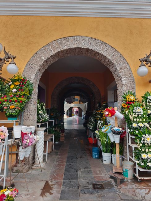 A storefront with an arched stone entryway features an arrangement of fresh flower bouquets and floral displays on both sides. The flowers include vibrant yellow, white, pink, and red blossoms, arrang