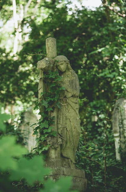 A weathered stone statue of an angel, draped in a flowing gown, leaning against a tall, narrow cross. The statue is covered partially in green ivy, which wraps around its neck and down its front, blen