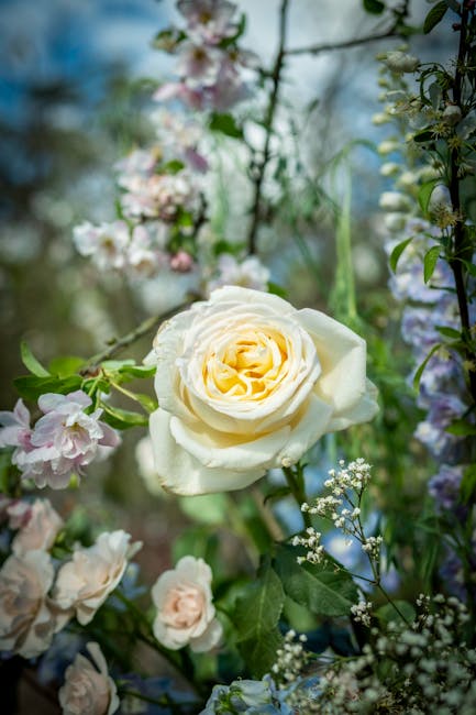 A close-up of a fresh, creamy white rose with delicate, layered petals, set among a variety of other flowers and greenery. Surrounding the rose are soft pink and white blossoms, small white filler flo